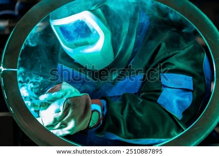 Welder intensely focused on welding a circular metal piece, with bright blue light reflecting off the protective mask.