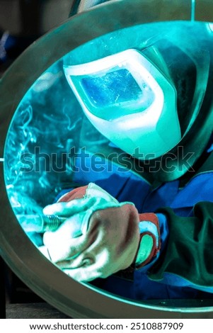 Welder intensely focused on welding a circular metal piece, with bright blue light reflecting off the protective mask.