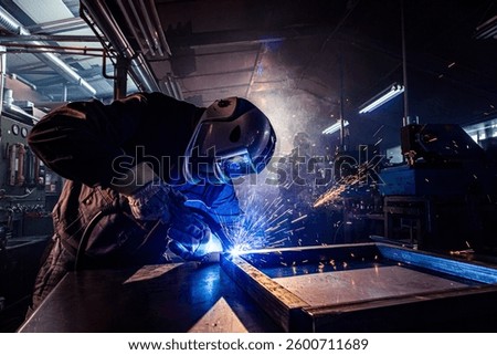 Welder joining metal structure using specialized equipment, creating sparks and smoke in industrial setting