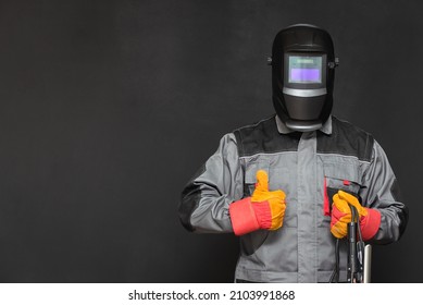 Welder man in the helmet and with welding terminals in the hand on the black wall background with copy space shows a thumbs up gesture. - Shutterstock ID 2103991868