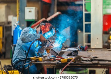 Welder performs arc welding on a metal workpiece at a workshop table, creating sparks during fabrication, repair and industrial metalworking operations.
