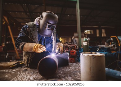 Welder in protective uniform and mask welding metal pipe on the industrial table while sparks flying.