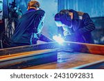 A welder wearing a protective mask for metal welding and protective gloves performs welding work at a metal structures plant. Welding a steel industrial beam. Sparks, hot flame from welding.