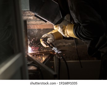 A Welder In A Welding Mask, Gloves Welds Metal With A Welding Machine And An Electrode In His Hand, Sparks Fly, A Beautiful Glow, Bokeh, Orange Light On A Black Background, A Flash Of Light And Smoke