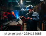 A welder is welding a metal part in car factory with the help of a robotic arm.