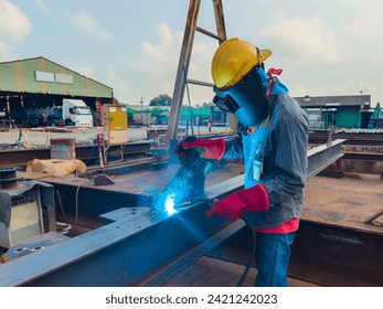 Welder Welding Steel Plate for Steel Structure Work with Process Flux Cored Arc Welding (FCAW) and Appropriately Dressed with Personal Protective Equipment (PPE) for Safety, at an Industrial Factory. - Powered by Shutterstock