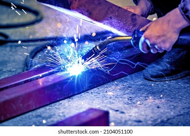 A welder welds metal into his workshop. Blue welding sparks. Gas combustion and blue smoke. Small welding workshop. Welding juncture of metal construction - Shutterstock ID 2104051100