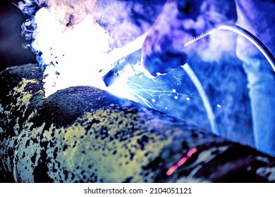 A welder welds metal into his workshop. Blue welding sparks. Gas combustion and blue smoke. Small welding workshop. Welding juncture of metal construction - Shutterstock ID 2104051121