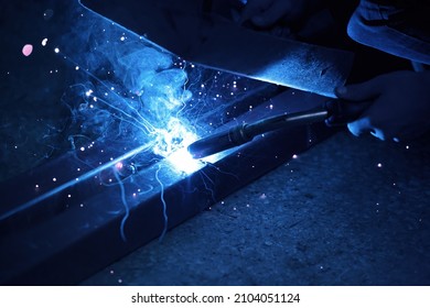 A welder welds metal into his workshop. Blue welding sparks. Gas combustion and blue smoke. Small welding workshop. Welding juncture of metal construction - Shutterstock ID 2104051124