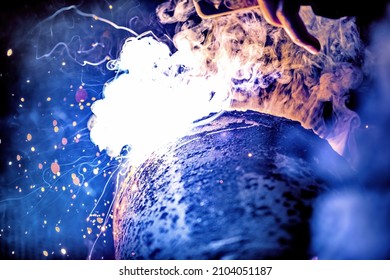 A welder welds metal into his workshop. Blue welding sparks. Gas combustion and blue smoke. Small welding workshop. Welding juncture of metal construction - Shutterstock ID 2104051187