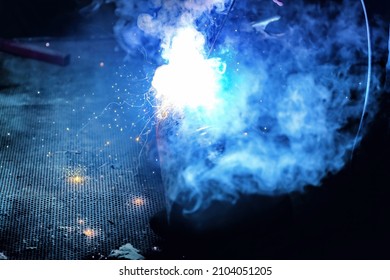 A welder welds metal into his workshop. Blue welding sparks. Gas combustion and blue smoke. Small welding workshop. Welding juncture of metal construction - Shutterstock ID 2104051205