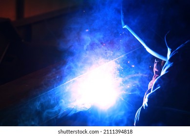 A welder welds metal into his workshop. Blue welding sparks. Gas combustion and blue smoke. Small welding workshop. Welding juncture of metal construction - Shutterstock ID 2111394818