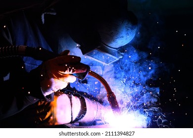 A welder welds metal into his workshop. Blue welding sparks. Gas combustion and blue smoke. Small welding workshop. Welding juncture of metal construction - Shutterstock ID 2111394827