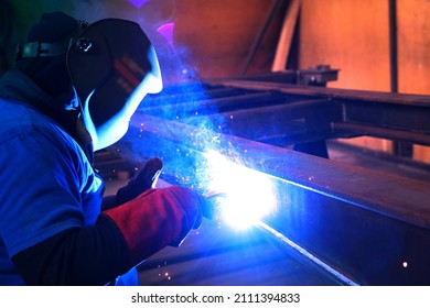 A welder welds metal into his workshop. Blue welding sparks. Gas combustion and blue smoke. Small welding workshop. Welding juncture of metal construction - Shutterstock ID 2111394833