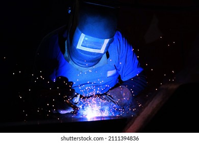A welder welds metal into his workshop. Blue welding sparks. Gas combustion and blue smoke. Small welding workshop. Welding juncture of metal construction - Shutterstock ID 2111394836