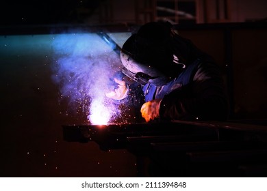 A welder welds metal into his workshop. Blue welding sparks. Gas combustion and blue smoke. Small welding workshop. Welding juncture of metal construction - Shutterstock ID 2111394848