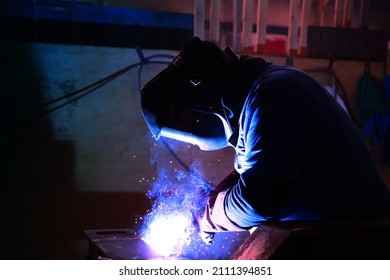 A welder welds metal into his workshop. Blue welding sparks. Gas combustion and blue smoke. Small welding workshop. Welding juncture of metal construction - Shutterstock ID 2111394851