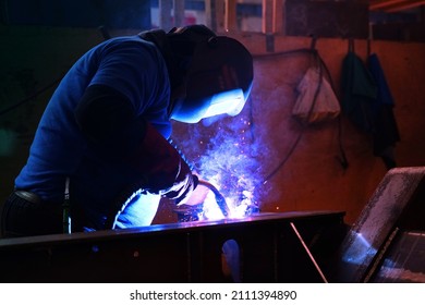 A welder welds metal into his workshop. Blue welding sparks. Gas combustion and blue smoke. Small welding workshop. Welding juncture of metal construction - Shutterstock ID 2111394890