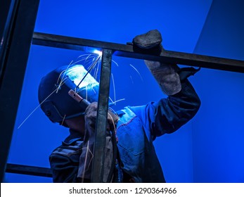 Welder Worker Performs Jump Welding. Worker Welder Performs Arc-Welding Process of Metal Structures. Flying Sparks From the Welding Machine. Welder Welds Metal Profiles for Second Floor Indoors