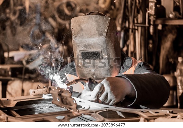 A welder worker in a protective metal mask is\
engaged in welding and repair of a car door, metal work in the\
workshop of the plant.