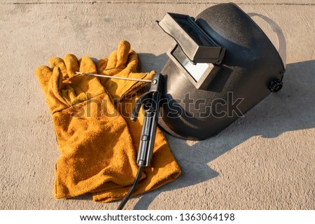 Welding equipment including leather welding gloves, welding mask, handheld welding electrode on concrete floor with orange sunlight. 