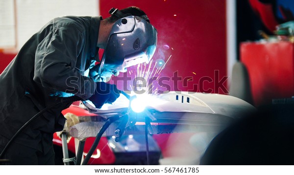 Welding industrial: worker in\
helmet repair detail in car auto service - blue sparklers,\
telephoto