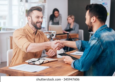 Well Done! Two Confident Young Men Shaking Hands And Smiling While Sitting At The Desk In Office With Two People Working In The Background 
