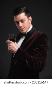 Well Dressed Good Looking Business Man Holding Glass Of Whiskey. Wearing Dark Red Dinner Jacket And Black Bow Tie.