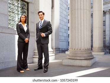 A Well Dressed Man And Woman Stand Among The Columns Of A Legal Or Municipal Building. Could Be Politicians, Business Or Legal Professionals.