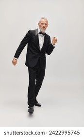well dressed mature man with stylish bow tie in voguish tuxedo dancing actively on gray backdrop