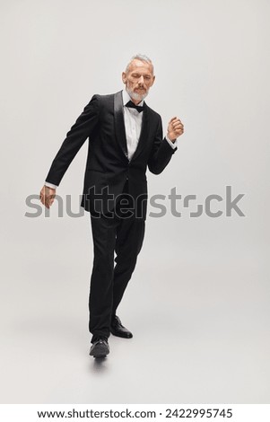 well dressed mature man with stylish bow tie in voguish tuxedo dancing actively on gray backdrop