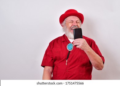 Well Dressed Old Man Wearing A Red Derby Hat, Red Shirt And Large Bolo Tie Making A E-commerce Purchase With His Smartphone.
Happy Old Man Is Having A Fun Conversation Over His Phone.