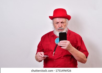 Well Dressed Old Man Wearing A Red Derby Hat, Red Shirt And Large Bolo Tie Making An E-commerce Purchase With His Smartphone.