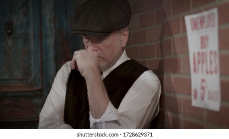 A Well Dressed Unemployed Man From The 1930s Great Depression Era Leaning Against A Wall Where A Sign Selling Apples For 5 Cents Each Is Posted.