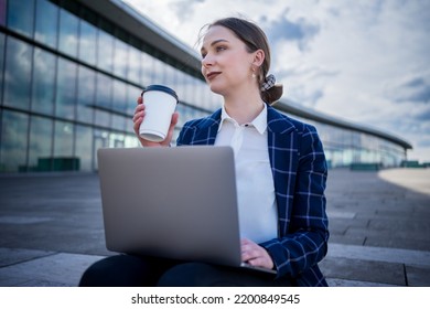 Well Dressed Young Businesswoman With Netbook And Cup Of Takeaway Coffee Looking Away While Sitting On Step And Working On Street Of Modern City