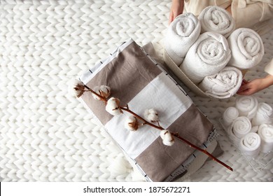 Well groomed woman hand holding a cotton branch with stack of neatly folded linens near rolled up towels in mesh basket placed on knitted chunky merino wool yarn plaid. Natural textile. Top view.