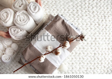 Well groomed woman hand holding a cotton branch with stack of neatly folded linens near rolled up towels in mesh basket placed on knitted chunky merino wool yarn plaid. Natural textile. Top view.