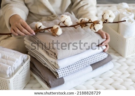 Well groomed woman hands holding the cotton branch with pile of neatly folded bed sheets, blankets and towels. Production of natural textile fibers. Manufacture. Organic product.