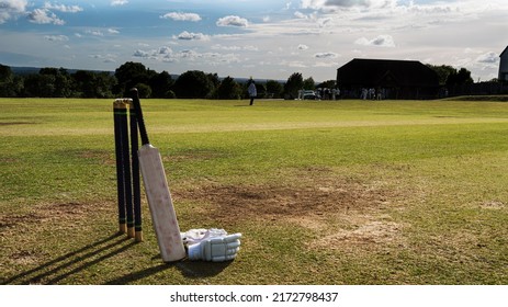 A well used cricket bat leaning against stumps in late evening sun, with umpire in background and batsman's gloves on ground - Powered by Shutterstock