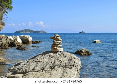 Well-balanced stack of rocks on a large, rough boulder on a beach on Skiathos island, Greece. Backdropped by the bright, calm aegean sea and a clear blue sky.