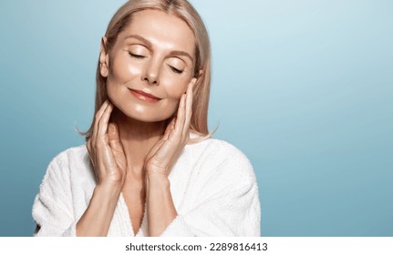 Wellbeing and beauty. Senior woman with perfect glowing skin, nourished face without blemishes, applies skincare routine, anti-aging cream for perfect glow and mousturizing effect, blue background. - Powered by Shutterstock