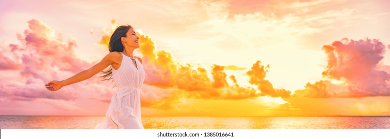 Wellbeing freedom happy woman jumping dancing of joy with open arms in the air blissful banner. Asian woman in sunset clouds pink sky background.