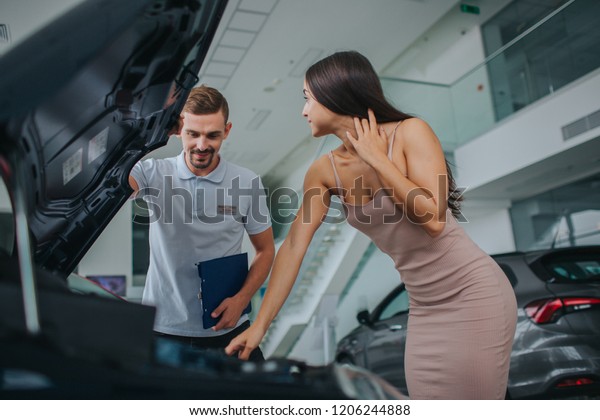 Well-built and nice brunette stands at opened body
of car and leans on it. She looks at consultant. He holds top body
of car and looks inside of it. Guy is concentrated. Young woman
touches her hair.