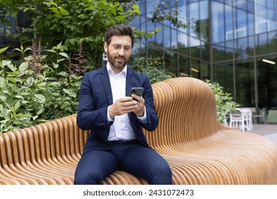 A well-dressed businessman engages with his smartphone on a modern wooden bench outside an office building. - Powered by Shutterstock