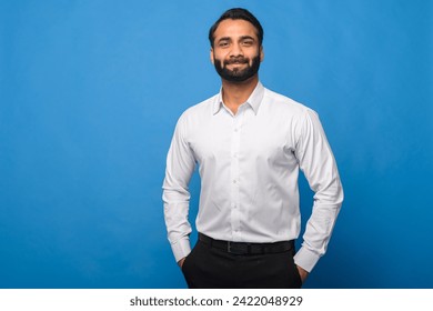 A well-dressed Indian man in a formal white shirt stands confidently with his hands in the pockets against a blue background. Professionalism, business, and smart casual attire. - Powered by Shutterstock