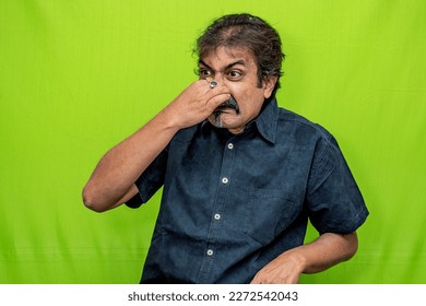 A well-dressed Indian man wearing a black shirt appears to be disgusted by a bad smell, as he holds his nose with his right hand while standing against a green screen background - Powered by Shutterstock