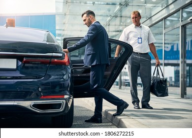 Well-dressed male traveler getting into a taxi while a porter holding his duffel bag standing by a car