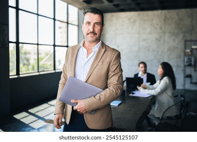 A well-dressed man holds a laptop in a stylish office filled with natural light. Colleagues engage in discussion at a nearby table, creating a dynamic work environment. - Powered by Shutterstock