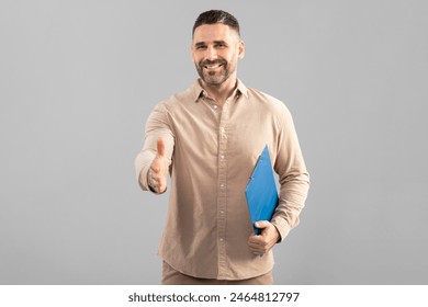 A well-dressed man smiles warmly as he extends his hand for a handshake. He holds a blue folder in his other hand, which may contain important documents. The background is plain and grey - Powered by Shutterstock