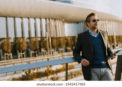 A well-dressed man smiles while using a tablet on a balcony, surrounded by contemporary architecture and vibrant autumn foliage, enjoying a moment of leisure. - Powered by Shutterstock